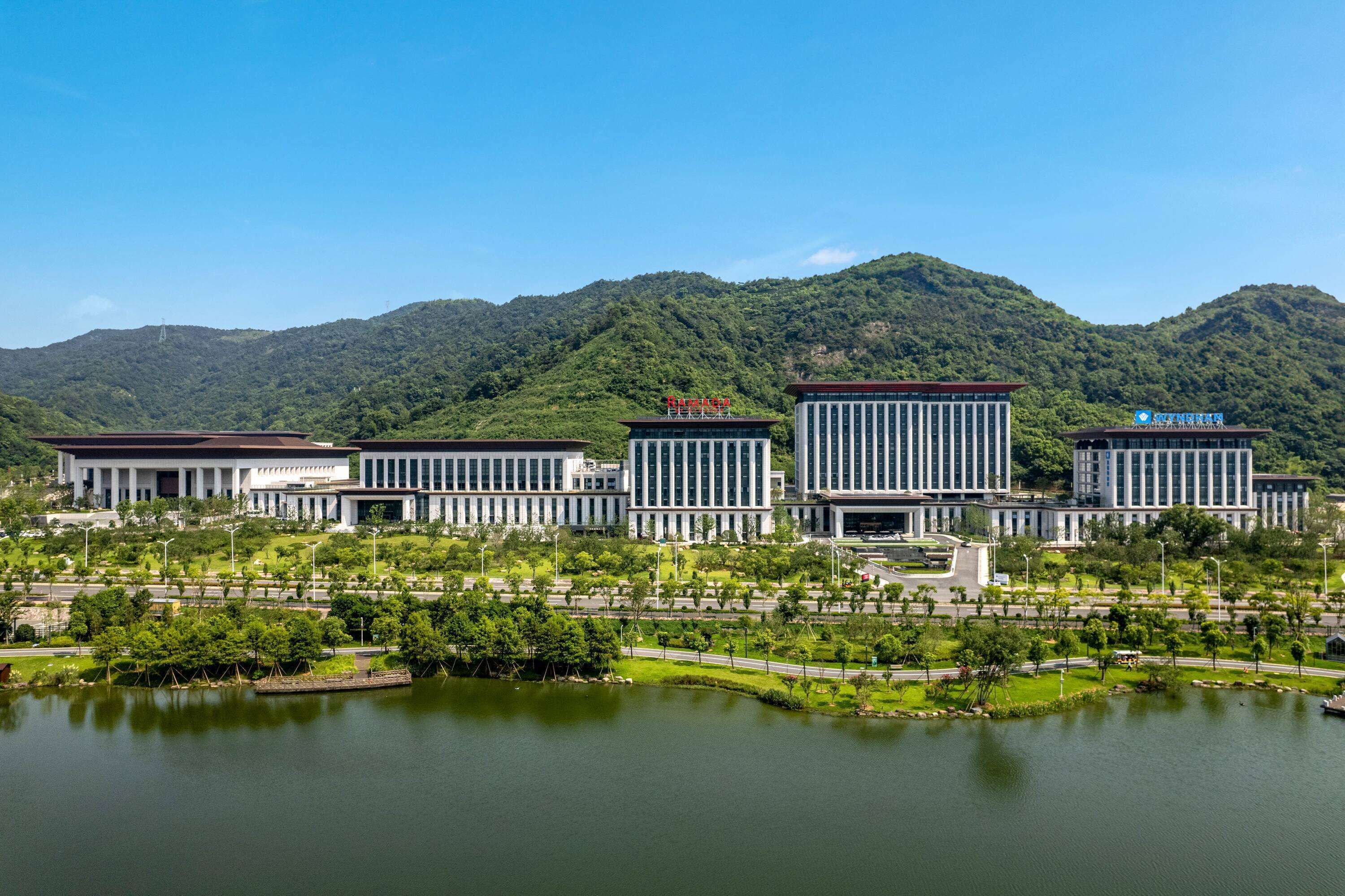A modern building complex with a central rectangular structure, surrounded by smaller buildings, set against a backdrop of lush green hills and a body of water.