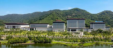A modern building complex with a central rectangular structure, surrounded by smaller buildings, set against a backdrop of lush green hills and a body of water.