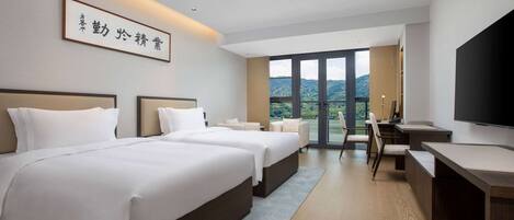 A hotel room with two beds, a desk, a chair, and a TV. There is a large window with a view of the outdoors.
