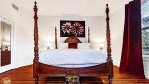 Een hemelbed met houten frame, een witte spreidek en een rood kussen. Boven het bed hangt een schilderij van bloemen op de muur.