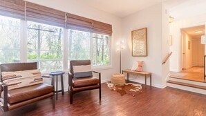 Un salón amplio con dos sillones de cuero, un otoman pequeño y redondo, y un banco con cojines. Ventanas grandes ofrecen luz natural y vistas al verdor exterior.