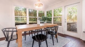 Un comedor con mesa de madera y sillas negras, ventanas amplias con vista a árboles, y una lámpara de araña.