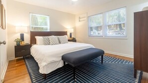 Un dormitorio con cama, mesitas de noche, un banco, ventilador de techo y ventanas.