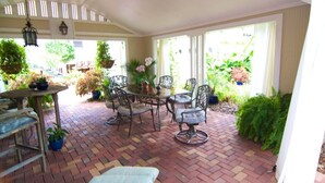 A covered patio area with a brick floor, metal chairs, a table, hanging lanterns, and potted plants.