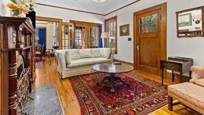 A spacious living room with a large sofa, a coffee table, and a patterned rug. There are wooden doors, a chandelier, and a small side table with a lamp.