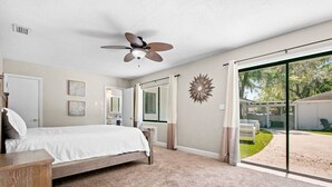 A bedroom with a bed, a ceiling fan, a large window, and a view of the outdoors.