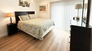 A bedroom with a bed, a nightstand, a lamp, a dresser, and a window with blinds. - 1 Bedroom Condo: Walk to Shands / UF / VA- Pool! (Gainesville)