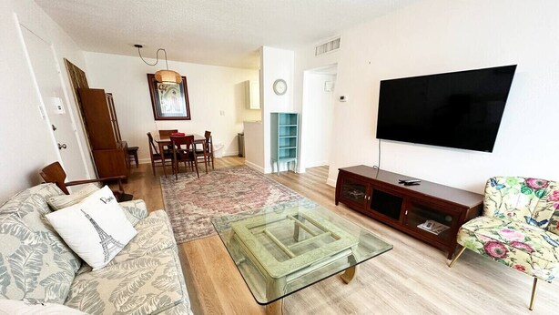 A living room with a glass coffee table, a sofa with patterned cushions, a floral armchair, a flat-screen TV, and a wooden cabinet. - 1 Bedroom Condo: Walk to Shands / UF / VA- Pool! (Gainesville)