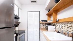 Une cuisine compacte équipée d’un réfrigérateur, d’un four, d’un four à micro-ondes et d’un évier. On trouve une étagère en bois au-dessus de l’évier et un pare-éclaboussures carrelé.
