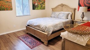 A bedroom with a wooden bed, a red and gold decorative basket, a nightstand with a lamp, and two wall-mounted paintings. - Large 1 BR Condo - Next to Oaks Mall and Hospital (Gainesville)