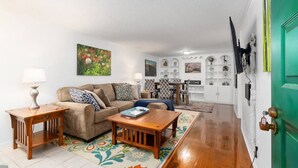 A living room with a sofa, coffee table, and side tables. There is a green door on the right and a white wall with framed pictures.