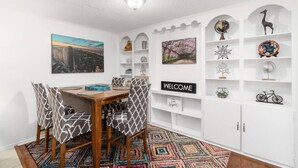 A dining area with a wooden table and patterned chairs, a colorful rug, and a 'WELCOME' sign on the wall.