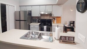 A compact kitchen with stainless steel appliances, a white countertop, and a sign that reads "KISS THE COOK". - Centrally located 2 bedroom condo for 6 people (Gainesville)
