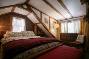 Quarto com teto inclinado, vigas de madeira, cama com roupa de cama floral, uma cadeira e uma escrivaninha.
