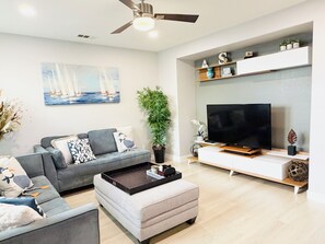Ein modernes Wohnzimmer mit einem grauen Sofa, einem weißen Hocker, einem Flachbildfernseher und einem Deckenventilator.