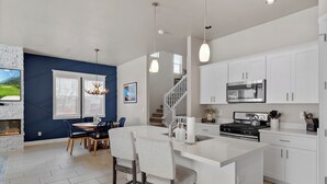 A modern kitchen with white cabinets, a central island, and a dining area with a blue wall and a fireplace. - Flagstaff Retreat w/ Hot Tub • Firepit • Dogs OK - 1 mi to NAU (Flagstaff)