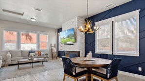 A modern living room with a fireplace, a dining area with a round table and chairs, and a large TV mounted on the wall. - Flagstaff Retreat w/ Hot Tub • Firepit • Dogs OK - 1 mi to NAU (Flagstaff)