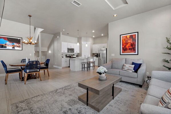 A modern living room with a dining area, a kitchen in the background, and a gray sofa with a painting.
