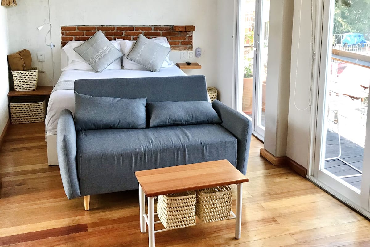 A bedroom with a bed, a grey sofa, a wooden table, and a view through a large window.