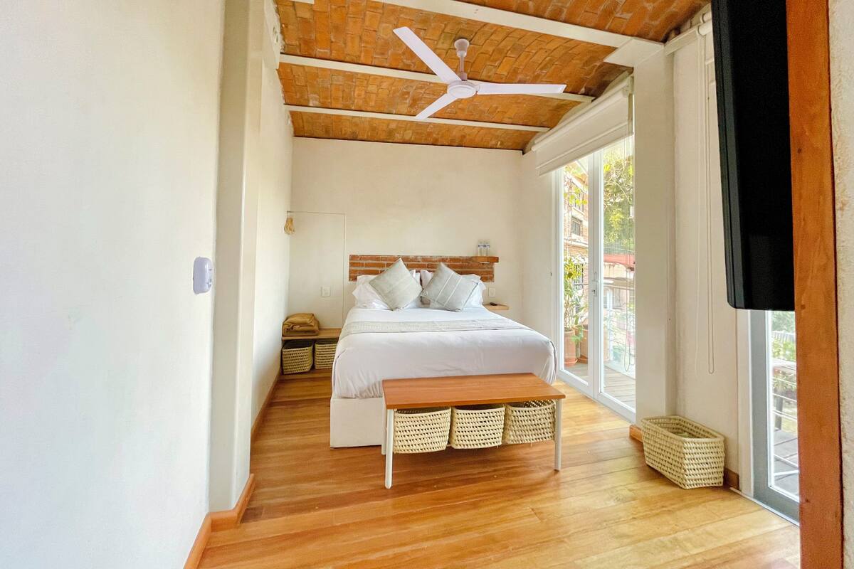 A bedroom with a wooden ceiling, a bed with white bedding, a ceiling fan, and a large window.