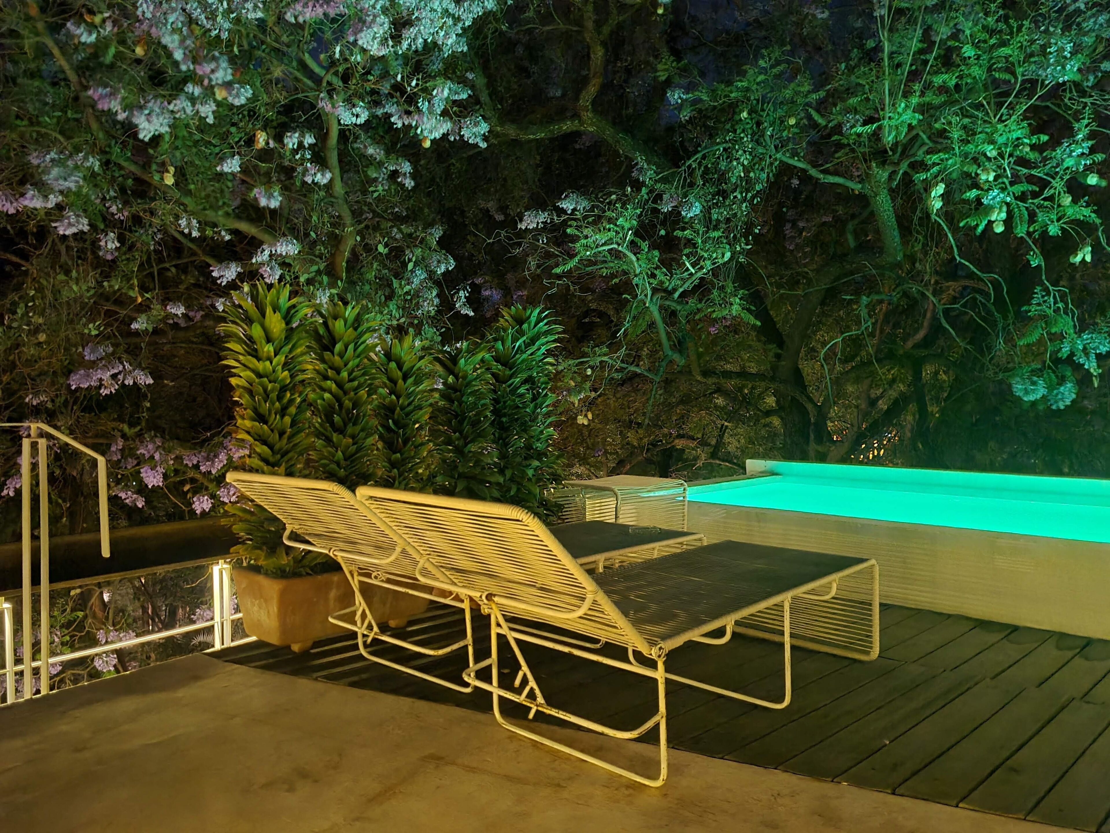 A deck with a metal chair and a table, surrounded by lush greenery and a pool.
