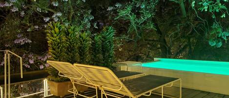 A deck with a metal chair and a table, surrounded by lush greenery and a pool.