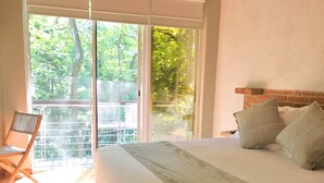 Un dormitorio con una cama grande, una silla de madera y vistas a la vegetación a través de un ventanal corredero.