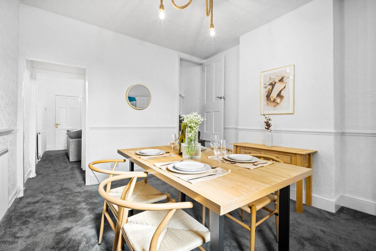 Ein Esszimmer mit einem Holztisch, gedeckt für vier Personen, umgeben von vier Korbstühlen. An der Wand hängt ein runder Spiegel, über dem Tisch befindet sich ein gerahmtes Kunstwerk und auf dem Tisch steht eine Vase mit Blumen.