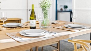 A dining table set with plates, cutlery, and a bottle of wine, with a vase of flowers and a clear glass container on the table. - Stylish 3 Bed House - 6 Guests - Garden - Parking (Birmingham)