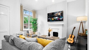 A living room with a grey sofa, a coffee table, a TV mounted on the wall, a fireplace, and a guitar. - Stylish 3 Bed House - 6 Guests - Garden - Parking (Birmingham)