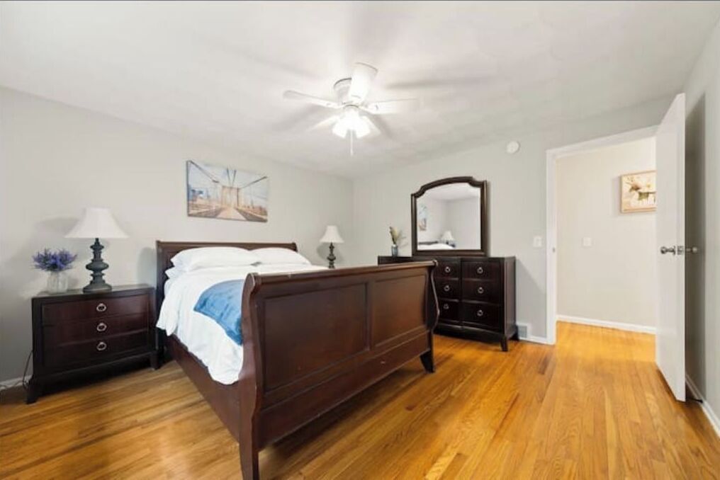 A bedroom with a wooden bed, a nightstand with a lamp, a dresser, a ceiling fan, and a door leading to another room.