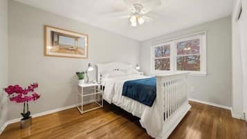 Une chambre avec un lit blanc, une table de chevet, un ventilateur de plafond, une fenêtre donnant sur un beau paysage et un tableau encadré accroché au mur.