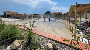 Een speelplaats met water, inclusief diverse waterstralen, omringd door een houten terras en een tegelpad.