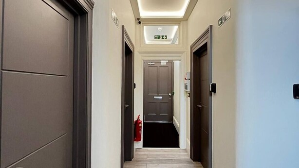 A modern hotel hallway with a central light fixture, a fire extinguisher, and a door with an emergency exit sign. - Apt. 04 -  Seaside Serenity Chic Studio Steps to Promenade (Brighton and Hove)