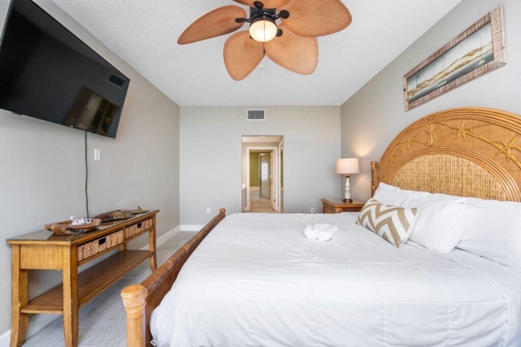 Un dormitorio con una cama grande, cabecera de madera, ventilador de techo, televisor montado en la pared y mesita de noche con lámpara.