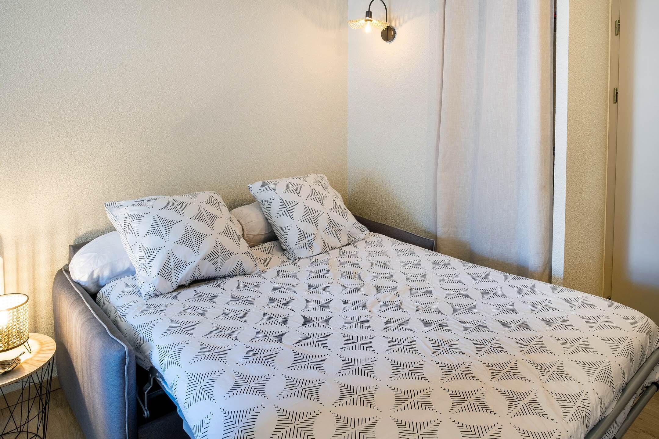 Un lit bien fait, avec une literie à motifs, une lampe de chevet et un luminaire fixé au mur.
