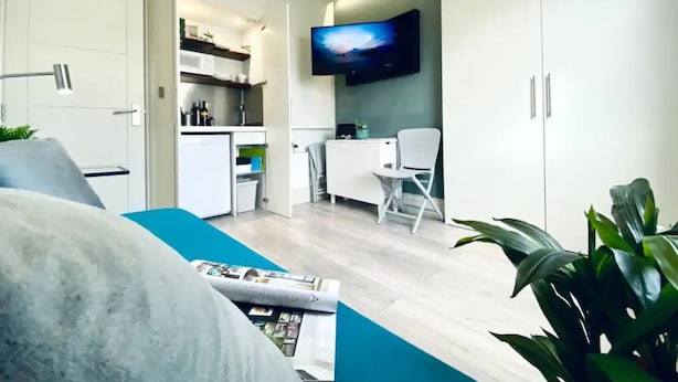 A modern living room with a sofa, a flat-screen TV mounted on the wall, a dining area with a table and chairs, and a kitchen area with cabinets and a microwave. - Apt. 05 - Stylish Studio Steps from Sea, Bars & Promenade (Brighton and Hove)