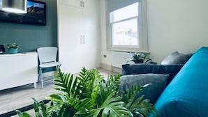 A modern living room with a white sofa, a blue sofa, a white desk, and a chair. There are potted plants and a large window with blinds. - Apt. 05 - Stylish Studio Steps from Sea, Bars & Promenade (Brighton and Hove)