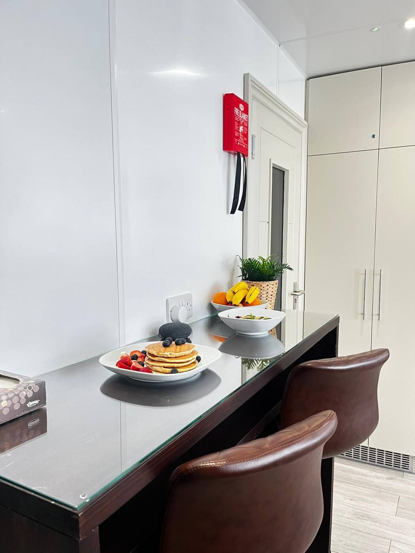 Een moderne keuken met een glazen aanrecht, een fruitschaal, een bord pannenkoeken en een rode brandblusser aan de muur.