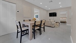 A modern living room with a dining area, a sofa, a TV mounted on the wall, and a door with louvered panels.
