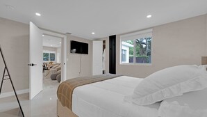 A modern bedroom with a large bed, a flat-screen TV mounted on the wall, and a window with a view of greenery outside.