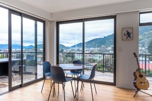 Um quarto espaçoso com janelas amplas, uma mesa redonda e cadeiras, um violão e um quadro na parede.