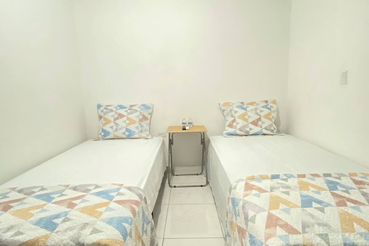 Une petite chambre propre, avec deux lits, chacun agrémenté d’une literie à motifs. Une petite table, sur laquelle se trouve une bouteille et une prise électrique, est placée entre les deux lits.