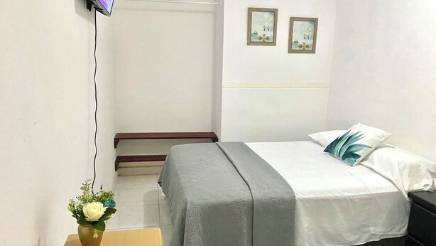 Un dormitorio con cama, mesita de noche, televisor montado en la pared y una pequeña mesa con un jarrón de flores.