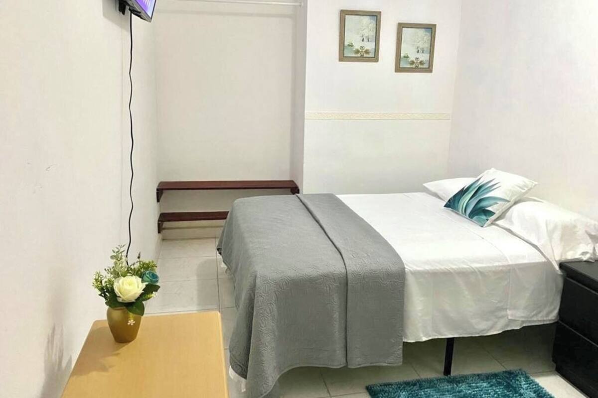 Une chambre à coucher avec un lit, une table de chevet, un téléviseur fixé au mur et une petite table sur laquelle est posé un vase de fleurs.