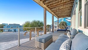 A covered outdoor deck with a wicker sofa and cushions, overlooking a street and buildings.
