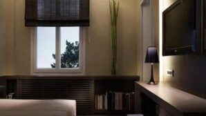 Une chambre d’hôtel avec un lit, une fenêtre avec des stores, une bibliothèque, un bureau avec une lampe et un téléviseur fixé au mur.