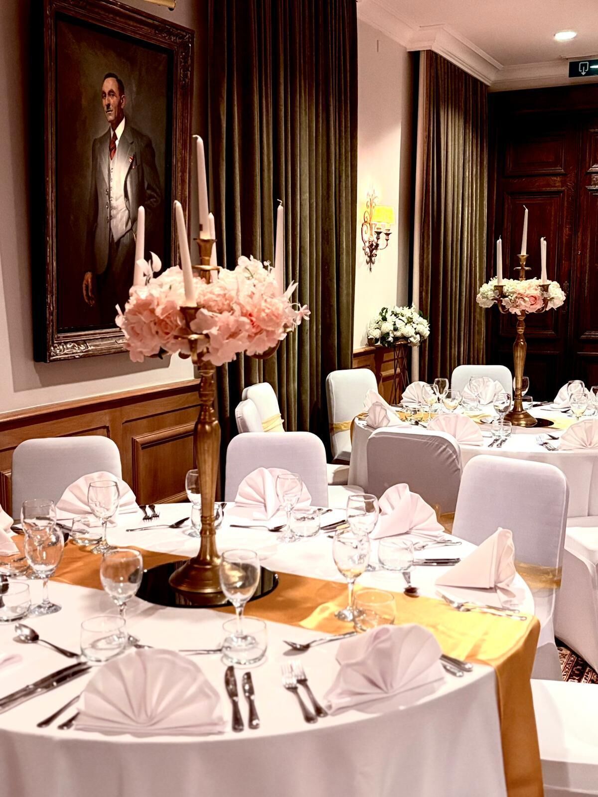 A formal dining room with round tables set for a meal, featuring white tablecloths, gold accents, and floral centerpieces.