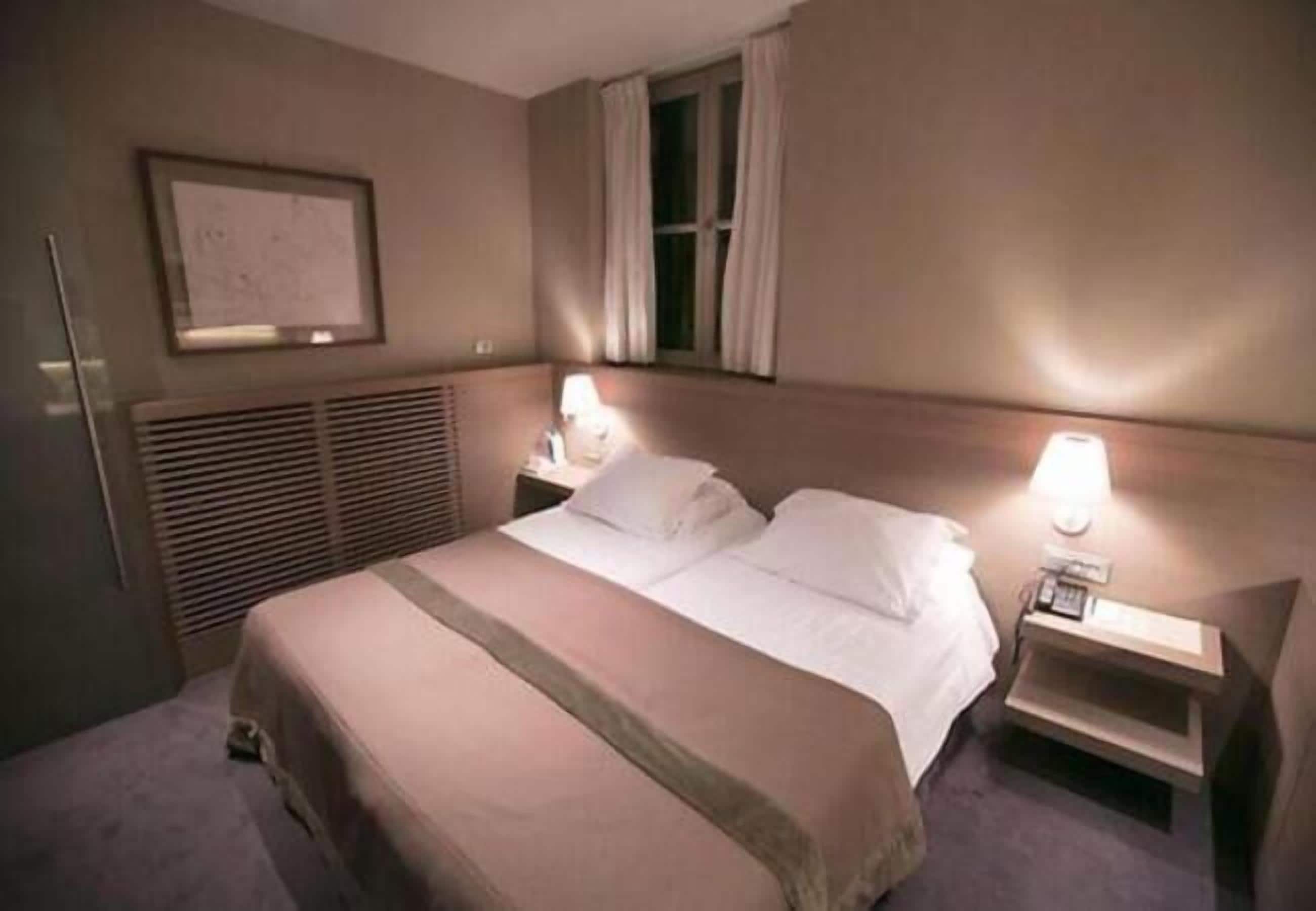 A hotel room with a bed, bedside tables, a window with curtains, a wall-mounted air vent, and a framed picture.