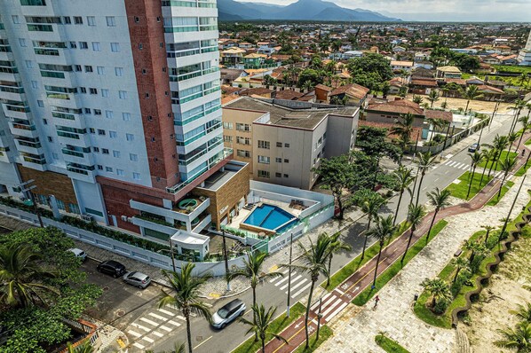 A high-rise building with multiple floors, a swimming pool, and a parking area with cars. - CANTINHO DA FAMÍLIA  (Praia Grande)